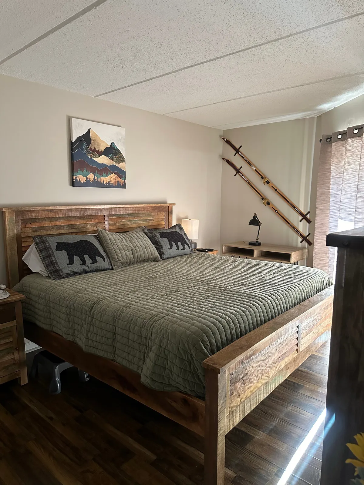 A spacious king suite featuring a large wooden bed adorned with bear motif pillows, mountain-themed artwork above the headboard, and rustic wooden furniture. Natural light gently streams in, creating a peaceful and inviting atmosphere perfectly suited for cozy mornings or restful evenings.