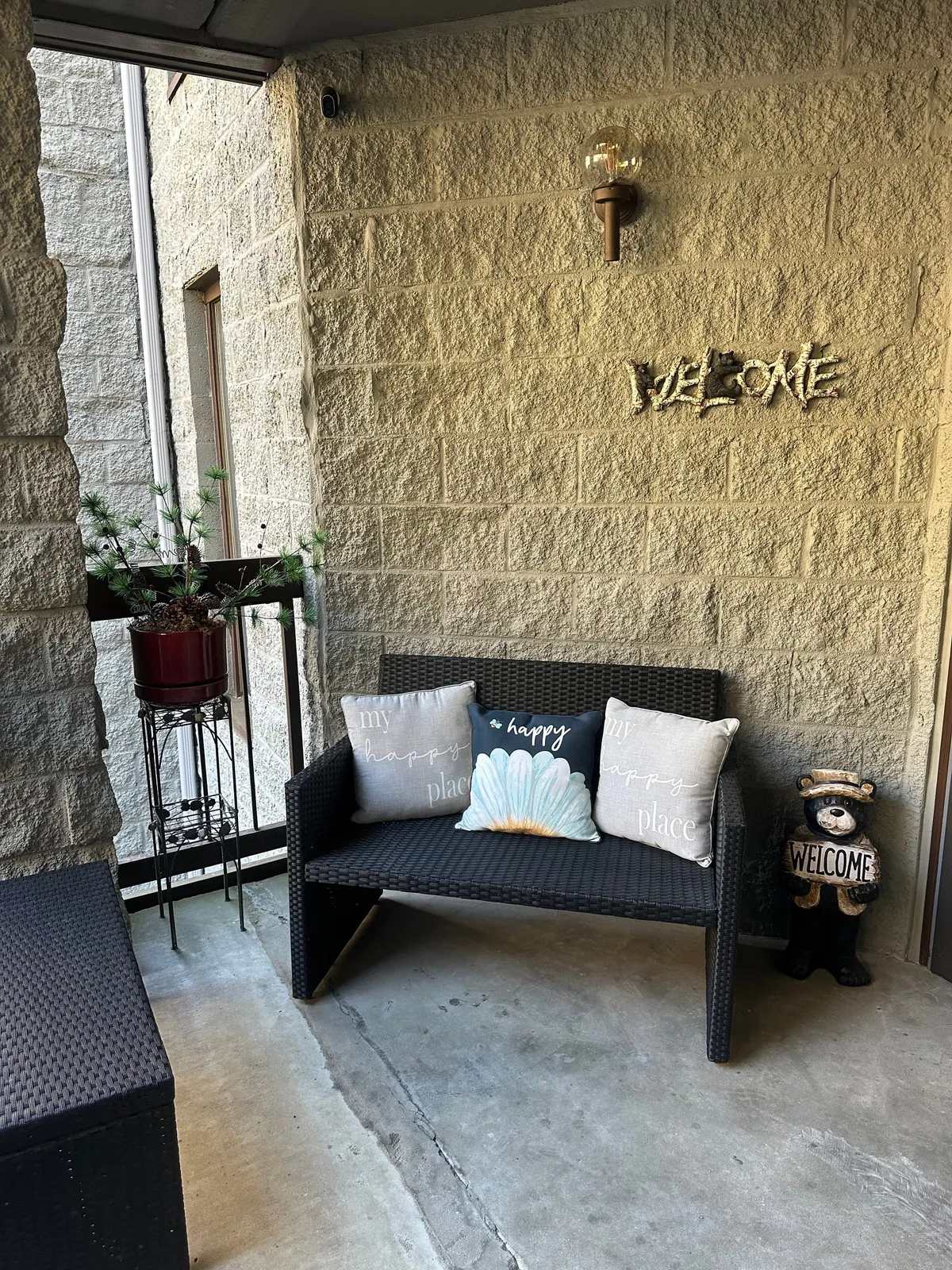 A cozy exterior porch scene featuring a wicker bench with cushions, a friendly welcome sign, a decorative bear statue, and potted plants. The space is ideal for relaxing or chatting after a day out, embodying warmth and hospitality.