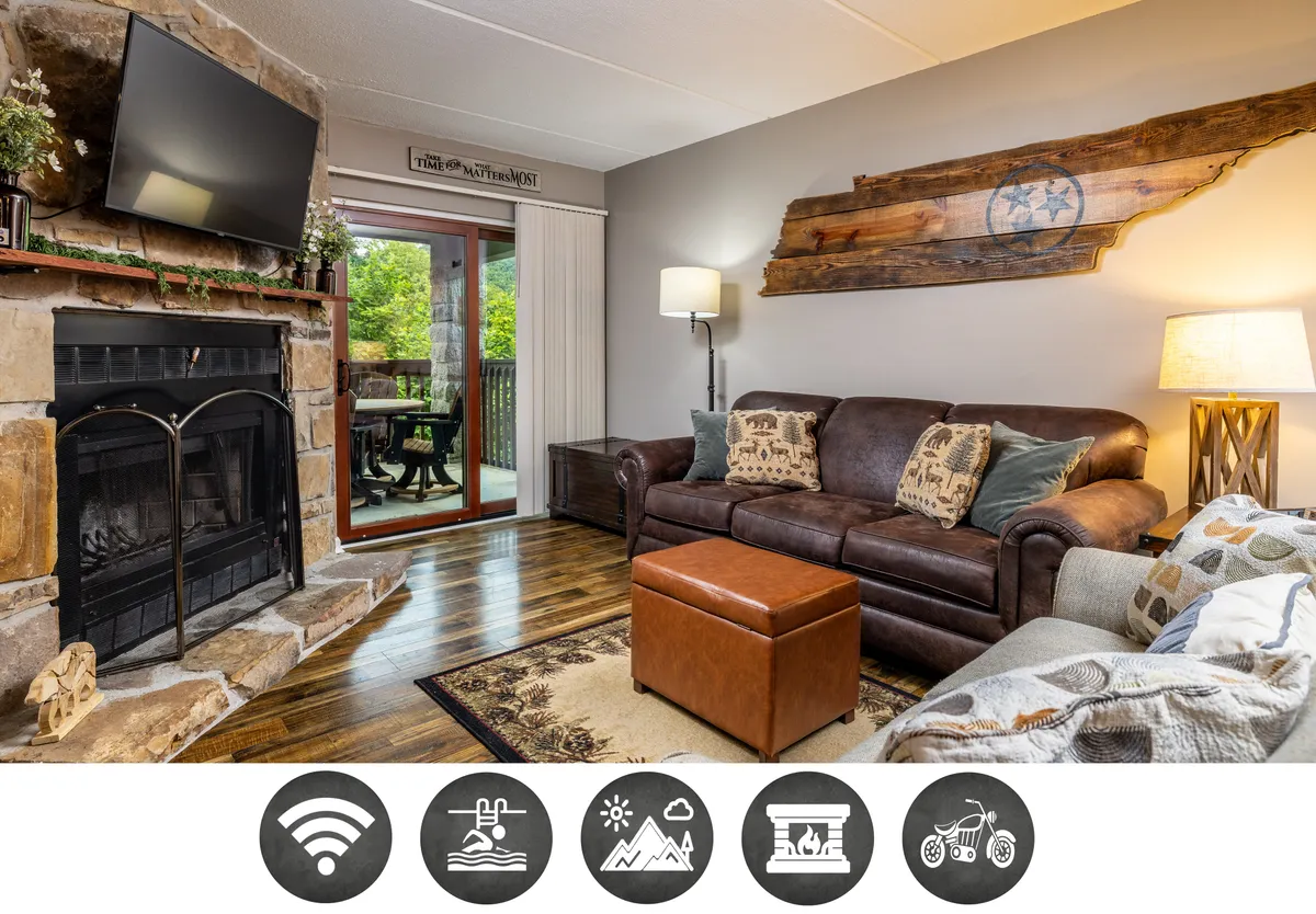 The image showcases a cozy and inviting living room with a crackling fireplace, plush sofa for lounging, rustic wooden wall art, and a sliding glass door leading to a balcony with a view—suggesting mountain vistas beyond. The warm lighting and thoughtful decor evoke a relaxed, welcoming atmosphere ideal for unwinding after a day of adventure.