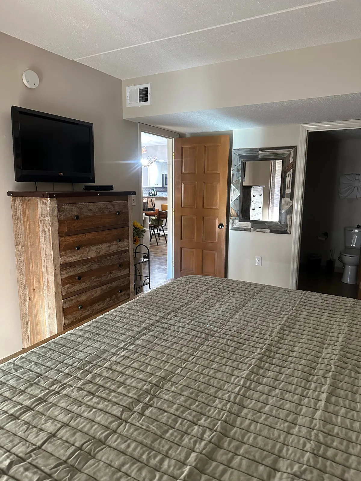 A cozy bedroom featuring a neatly made bed with a quilted cover, a wooden dresser, a television, and a doorway that opens toward the kitchen area. The overall mood is inviting and comfortable, suggesting a personal retreat for relaxation or unwinding with entertainment.