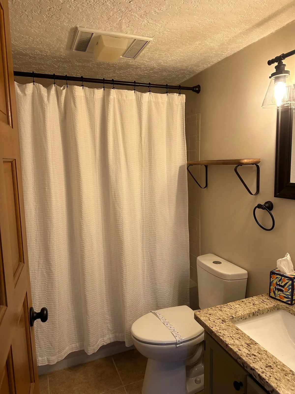 A bright bathroom with a patterned shower curtain, vanity with sink, wall-mounted shelf, and modern light fixture. The space looks clean and comfortable, perfect for getting ready before heading out or winding down at night.