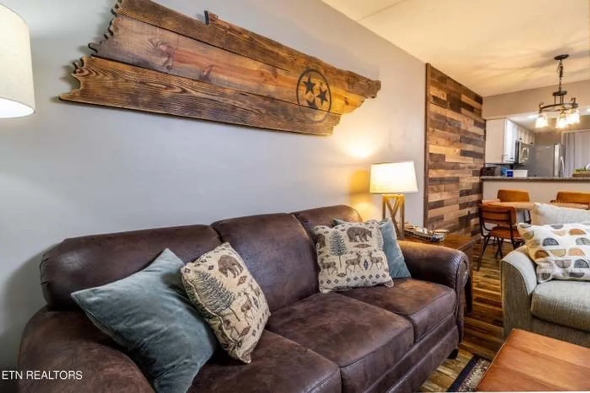 A cozy living area anchored by a leather couch with decorative pillows, set against a wooden accent wall featuring a wooden Tennessee state art piece. A table lamp casts a warm glow, creating an inviting space ideal for relaxing, socializing, or unwinding by the fire.