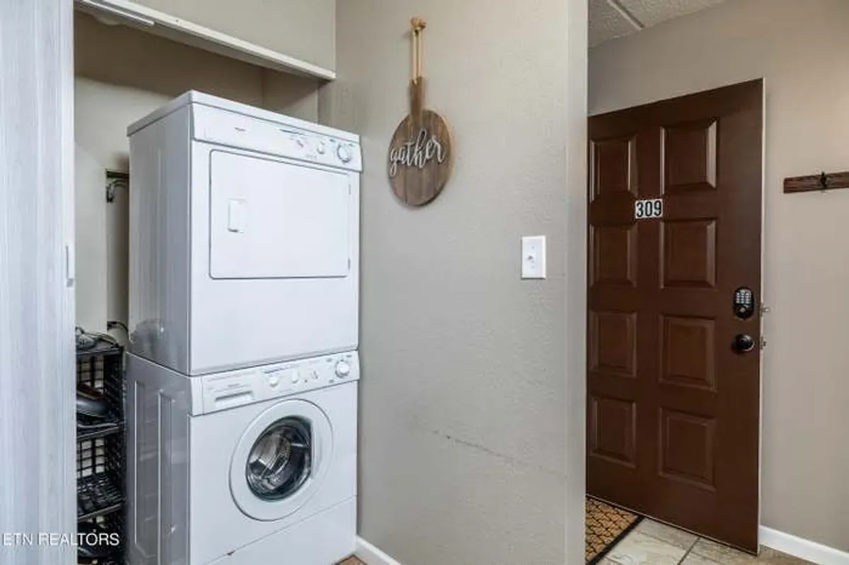 This image features a stacked washer and dryer beside a wooden 'gather' sign and the front door, situated in a nook that feels both practical and welcoming. The proximity to the entrance suggests ease of access for guests returning from outdoor adventures who need to clean up or drop off muddy gear.