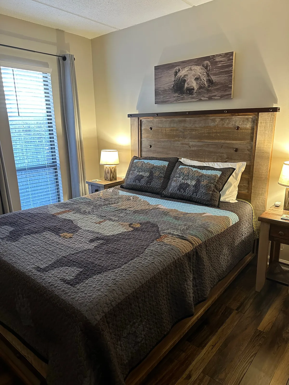 A warm and inviting bedroom with a bed adorned in a bear-themed quilt, a rustic wooden headboard, table lamps, and a photograph of a bear above the bed. Natural light softly fills the space, creating a peaceful environment for rest.