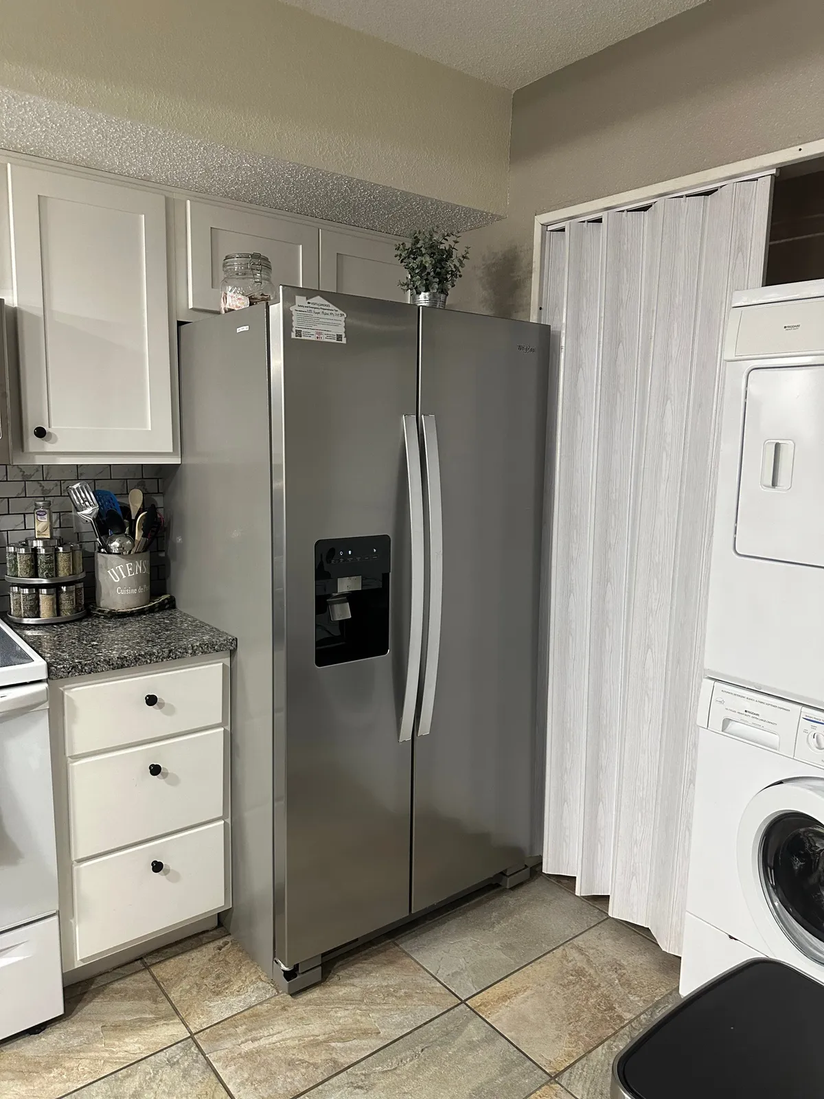 A practical, well-lit view of the kitchen’s stainless steel refrigerator, white cabinetry, granite countertop, and visible laundry appliances. The image combines meal prep and laundry areas, underscoring convenience and modern amenities.