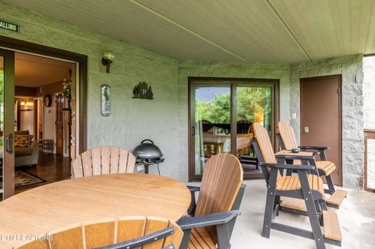 A cozy outdoor patio with a round wooden table surrounded by Adirondack chairs, a barbecue grill, and a backdrop of natural greenery. The setting is perfect for outdoor dining, relaxing, or enjoying a morning coffee while soaking in mountain air.