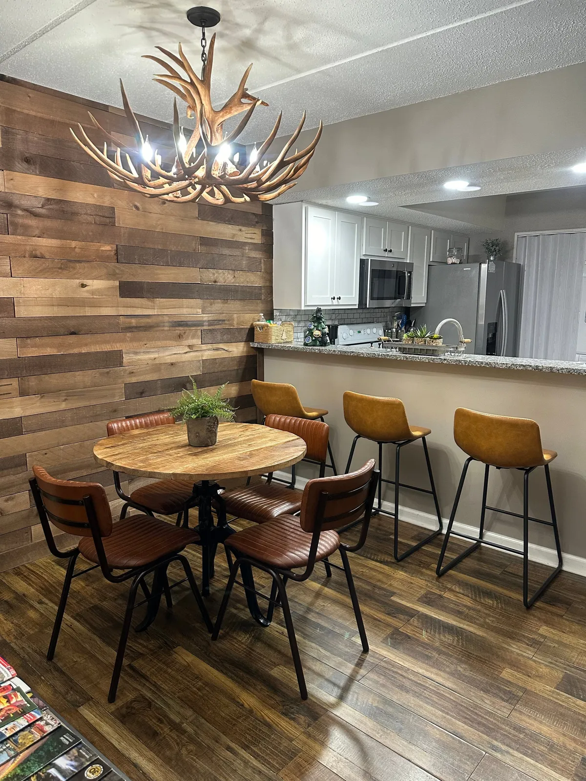 The image shows a warm, open dining area with a large wooden table, antler chandelier, and bar stools. The setup suggests a space for both intimate breakfasts and festive gatherings, tying together the kitchen and dining experience.