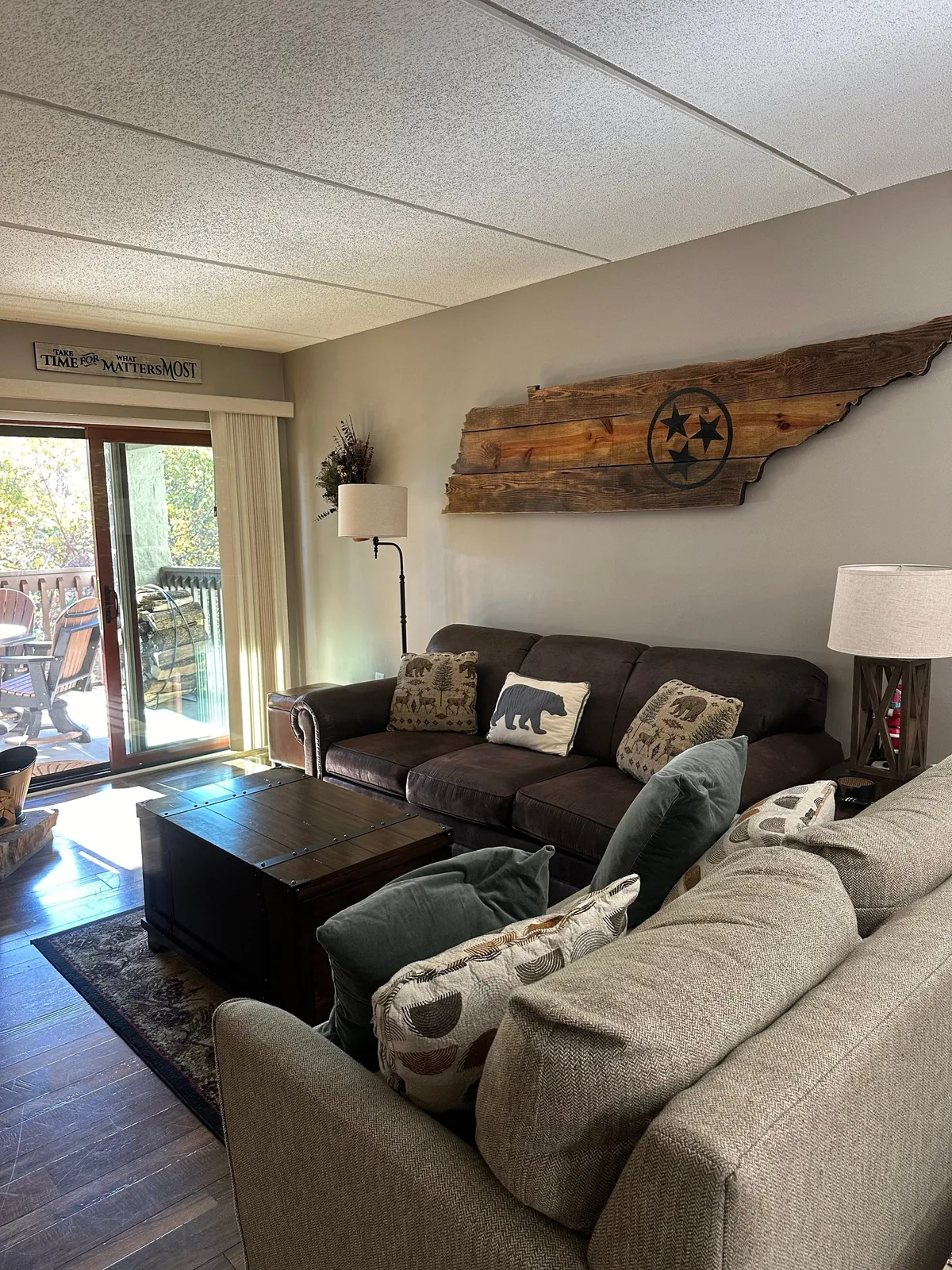 A spacious and inviting living room with a large sectional sofa, wooden coffee table, Tennessee-shaped wood decor on the wall, animal print pillows, and sliding glass doors opening directly to a private balcony. The space is illuminated by natural light and exudes warmth and comfort.
